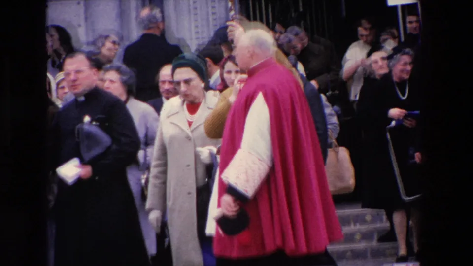 A red-caped clergyman appears near a stone entrance in the 1969 Paris street scene, surrounded by onlookers and another dark-robed religious figure.