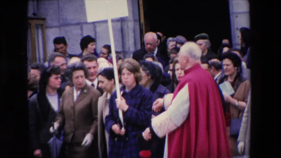 The red-caped clergyman is seen from behind as he moves through the Paris crowd, with a tall placard and closely packed onlookers in period clothing.