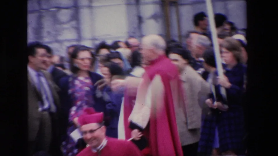 A soft, handheld 8mm frame shows the red-caped clergyman amid a crowded Paris public gathering, with a raised placard and onlookers filling the background.