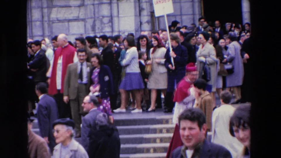 At the end of the clip, the crowd continues moving down the Paris church steps, with clergy in red vestments, a Landes placard, and several close foreground faces visible.