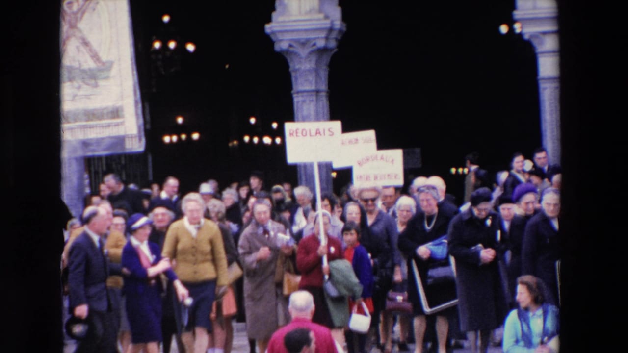 Paris, 1969 home-movie frame showing a crowd in coats and hats leaving a grand arched building while several people carry French-language placards.
