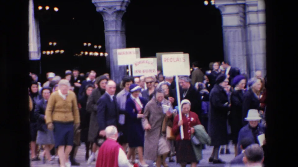 Paris, 1969 home-movie still with a moving crowd on stone steps, French-language signs overhead, and people in late-1960s coats, hats, and handbags.