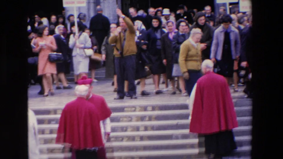 Paris, 1969 frame showing people dispersing down broad steps as a red-robed clerical figure stands in the foreground and signs remain visible within the crowd.