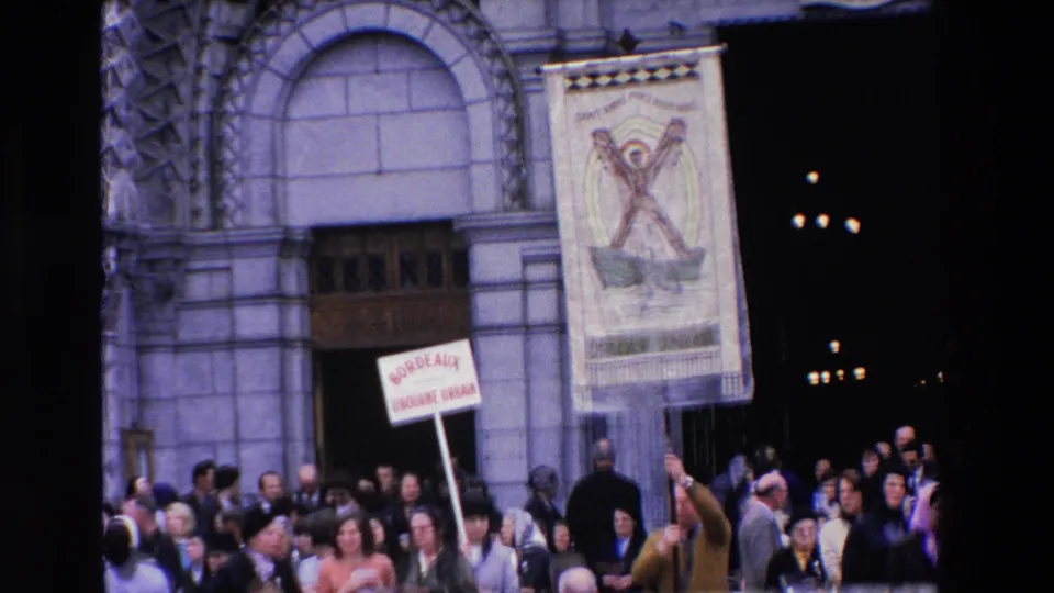 1969 Paris frame showing spectators beneath a stone facade and arched doorway, with a raised placard that appears to reference Bordeaux and Romainville.