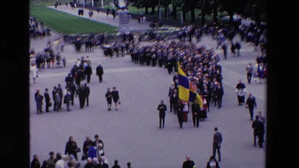Wide 1969 Paris home-movie view of an organized procession crossing a public square, with spectators lining the route and a colorful flag near the front.