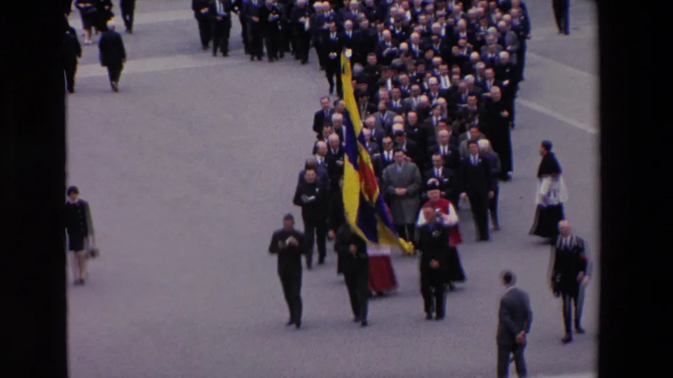 Elevated 1969 Paris home-movie view of a formal religious procession, with suited men and clergy walking behind a large yellow and blue flag.