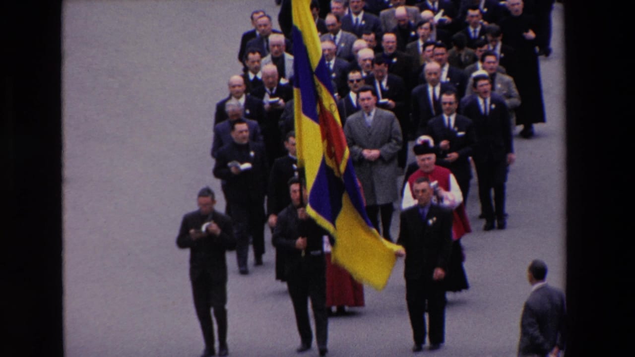 Closer view of a flag bearer and suited men walking in a formal street procession.
