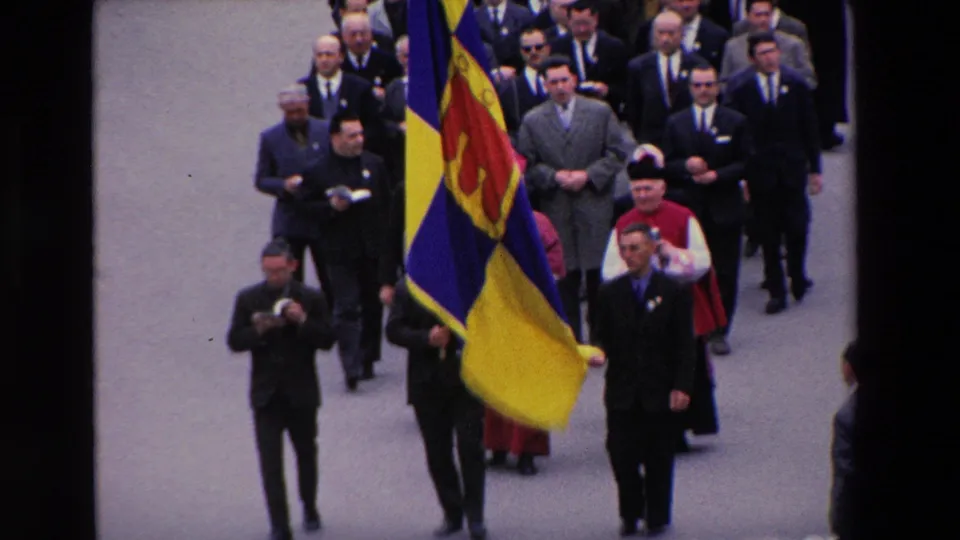 1969 Paris procession frame showing suited participants and clergy moving in close formation behind a prominent yellow and blue ceremonial flag.