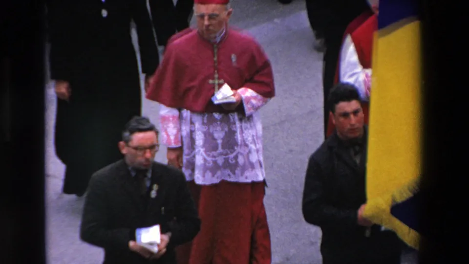 Close 1969 Paris home-movie frame of a clergy member in red and white vestments walking in the religious procession near the large flag.