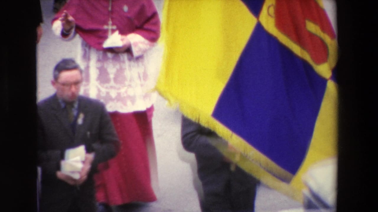 Paris 1969 home-movie frame showing a bishop in magenta vestments and clergy walking in a street procession, partly obscured by a large yellow and blue flag.