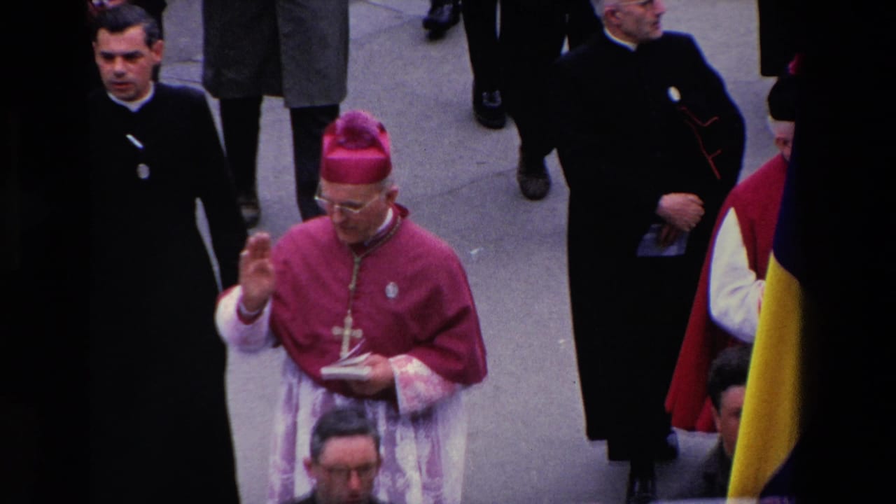 Bishop in magenta vestments walks with clergy and raises one hand during the procession.