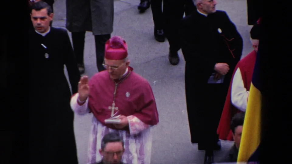 Paris 1969 home-movie frame showing a bishop in magenta vestments holding a small book or booklet and raising his hand while clergy and suited men walk nearby.