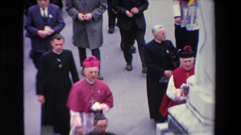 Paris 1969 home-movie frame showing the bishop in magenta vestments, nearby clergy, suited men, and a partially visible robed figure as the procession passes along the street.