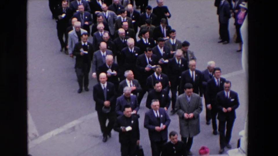 High-angle 1969 Paris 8mm frame showing rows of men in suits and ties walking in a formal street procession, with some carrying small papers.