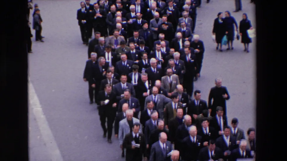 1969 Paris home-movie frame showing a tightly packed group of men in suits walking in formation, with several observers and women in dark clothing at the side.