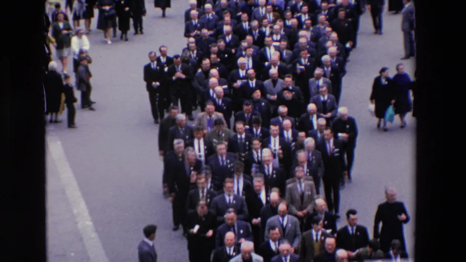 High-angle 1969 Paris frame showing a broad formal procession of men in suits, including possible clergy, moving along a city street with bystanders nearby.