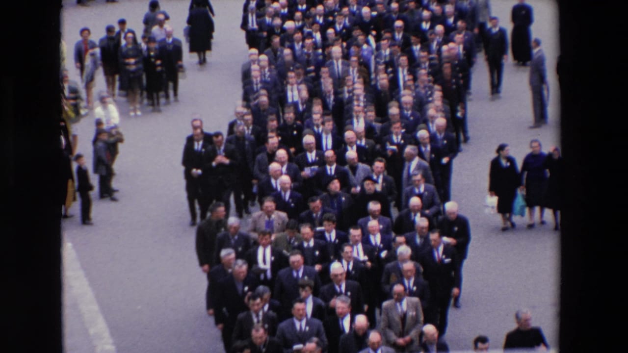 High-angle 1969 Paris home-movie frame showing rows of men in dark suits and ties moving forward in a formal street procession, with bystanders at the edges.