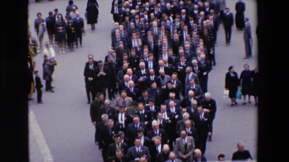 High-angle 1969 Paris home-movie frame showing rows of men in dark suits and ties moving forward in a formal street procession, with bystanders at the edges.