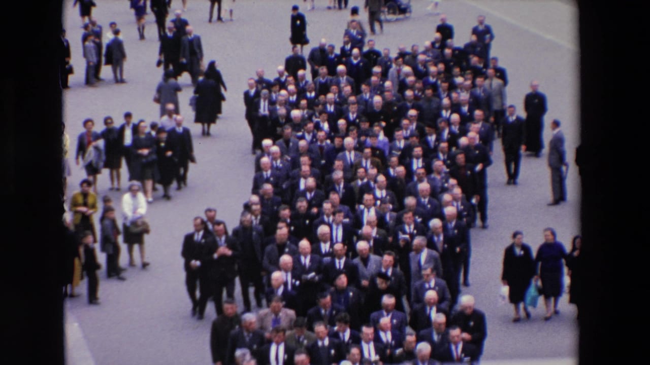 Dense procession of suited men with spectators standing around the street.