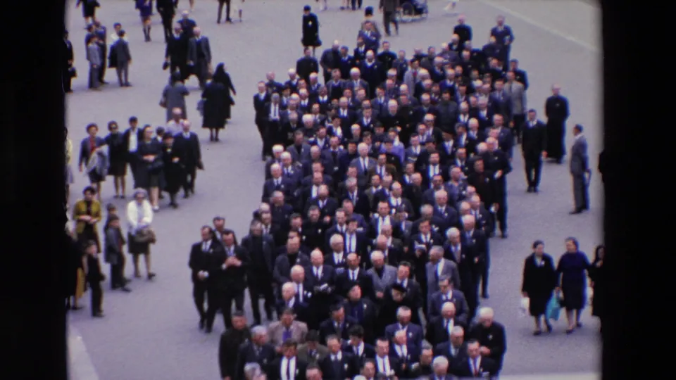 High-angle 1969 Paris frame of a dense formal procession of men in suits, surrounded by scattered spectators and a few women in dark coats.