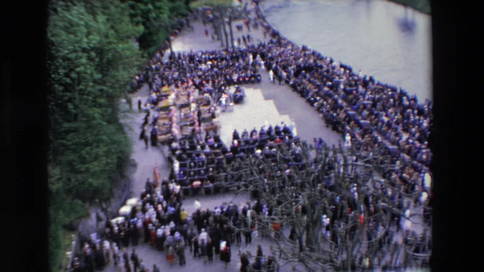 High-angle 1969 Paris frame showing a large formal assembly with rows of seated people, a surrounding crowd, bare tree branches, and greenery beside the gathering area.