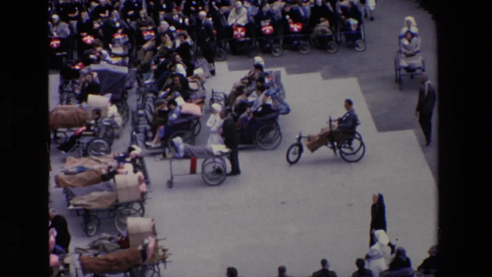 High-angle 1969 Paris home-movie frame showing a crowded plaza with wheelchair users, blanket-covered stretchers, nurses, and attendants.