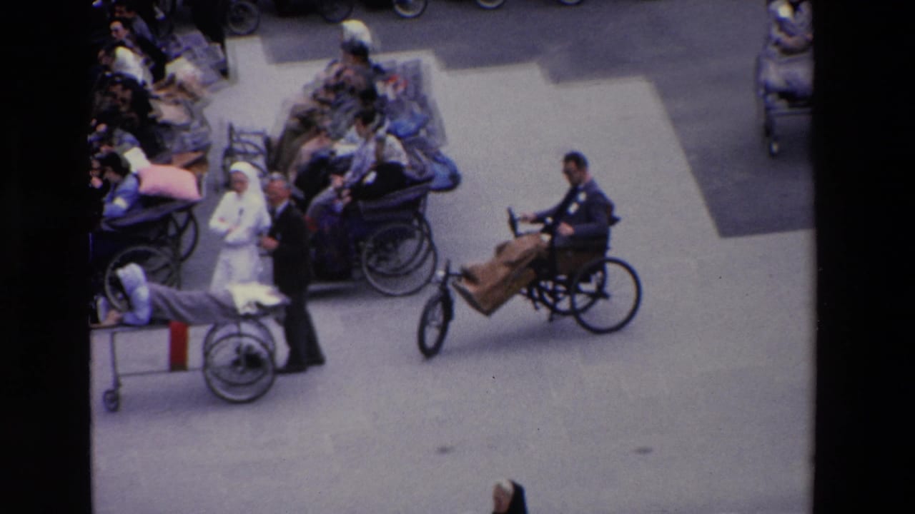 1969 Paris frame showing a wheelchair user with a wooden front attachment passing through a plaza crowded with nurses, attendants, and seated people.
