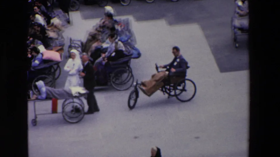 1969 Paris frame showing a wheelchair user with a wooden front attachment passing through a plaza crowded with nurses, attendants, and seated people.