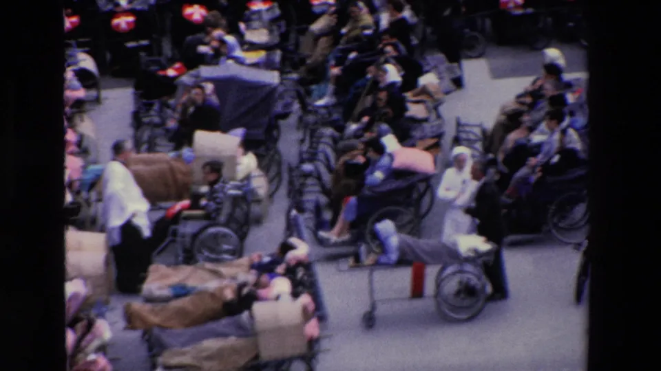 High-angle 1969 Paris frame with nurses in white moving among wheelchair users, stretchers, blankets, and seated onlookers in a public plaza.