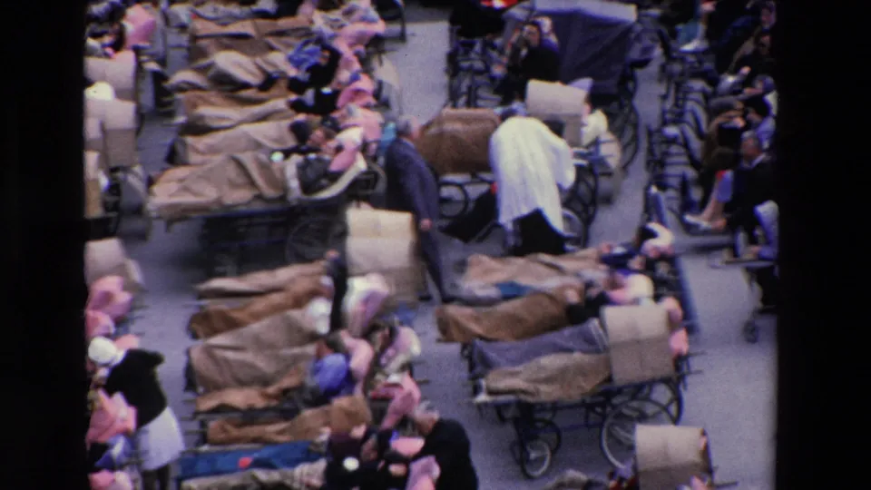 1969 Paris home-movie frame showing multiple wheeled stretchers with blanket-covered people, surrounding wheelchairs, and attendants moving through the crowded plaza.