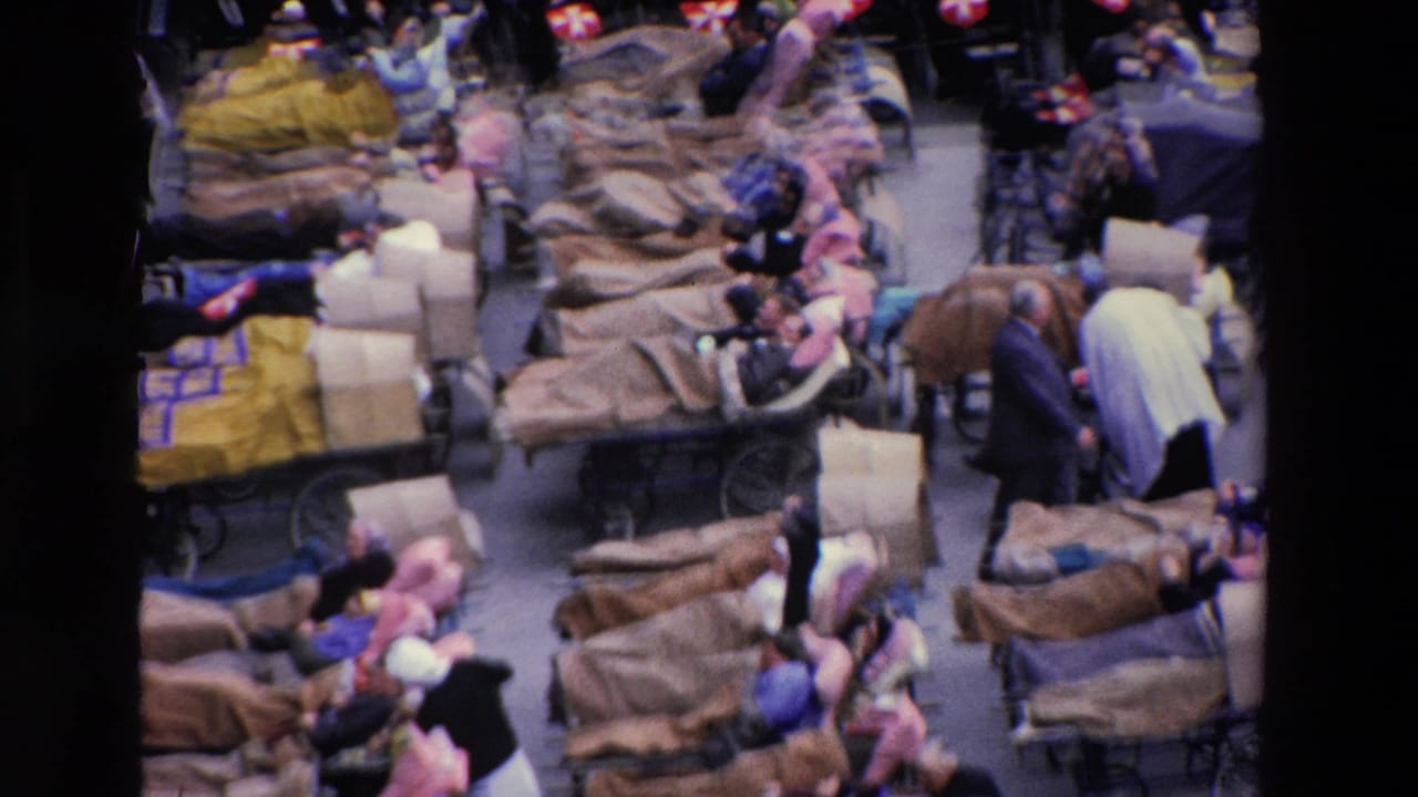 1969 Paris home-movie frame showing a crowded medical or emergency care scene with blanket-covered stretchers, reclining adults, attendants, and dense surrounding foot traffic.