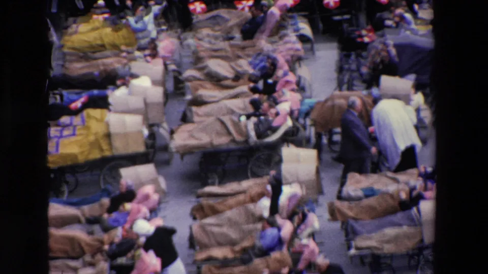 1969 Paris home-movie frame showing a crowded medical or emergency care scene with blanket-covered stretchers, reclining adults, attendants, and dense surrounding foot traffic.