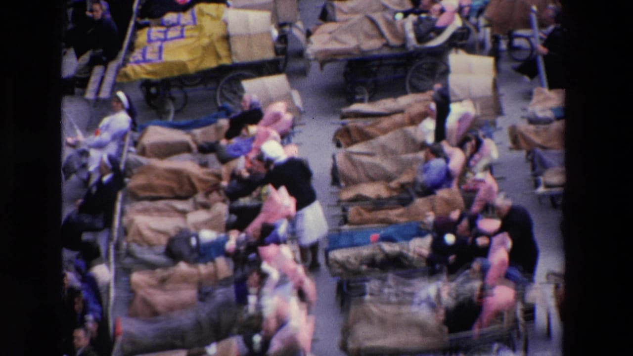 Rows of stretchers and seated people fill a crowded space viewed from above.