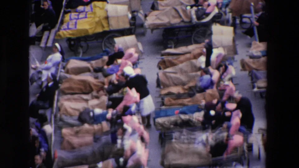 Elevated 8mm frame from Paris in 1969 showing rows of wheeled stretchers, blanket-covered people, seated adults, and attendants in a packed care area.