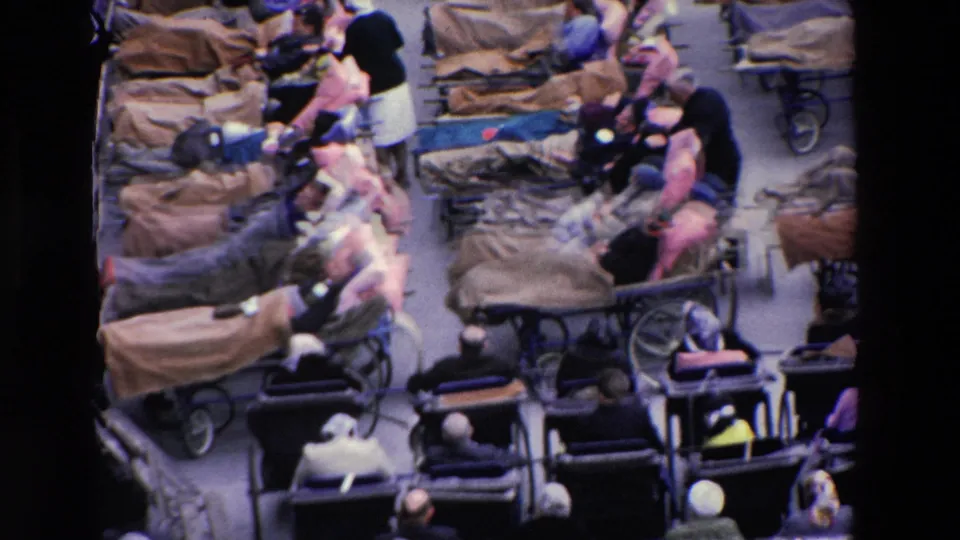 Closer 1969 Paris frame showing adults on blanket-covered gurneys, surrounding chairs, nearby attendants, and a tightly packed medical or emergency setting.