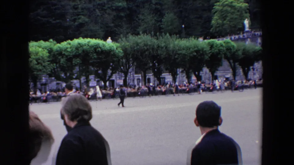 1969 Paris home-movie frame showing a broad paved public square, two foreground onlookers, distant gathered spectators, clipped trees, and a small statue-like figure in the distance.