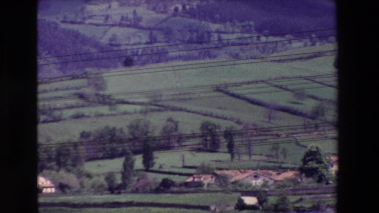 1960s 8mm home-movie frame of a rural landscape, possibly Spain-associated by metadata, with patchwork fields, scattered trees, small buildings, and power lines in the foreground.