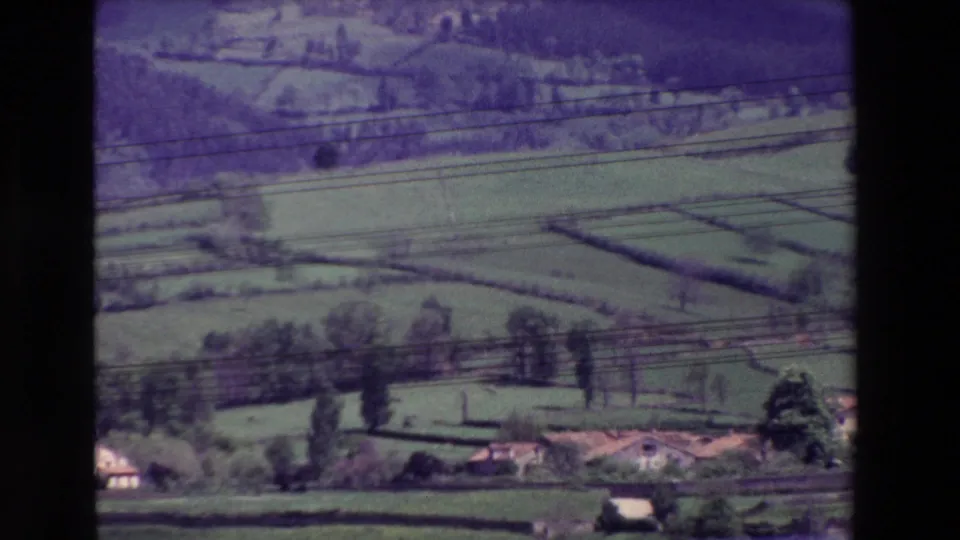 1960s 8mm home-movie frame of a rural landscape, possibly Spain-associated by metadata, with patchwork fields, scattered trees, small buildings, and power lines in the foreground.