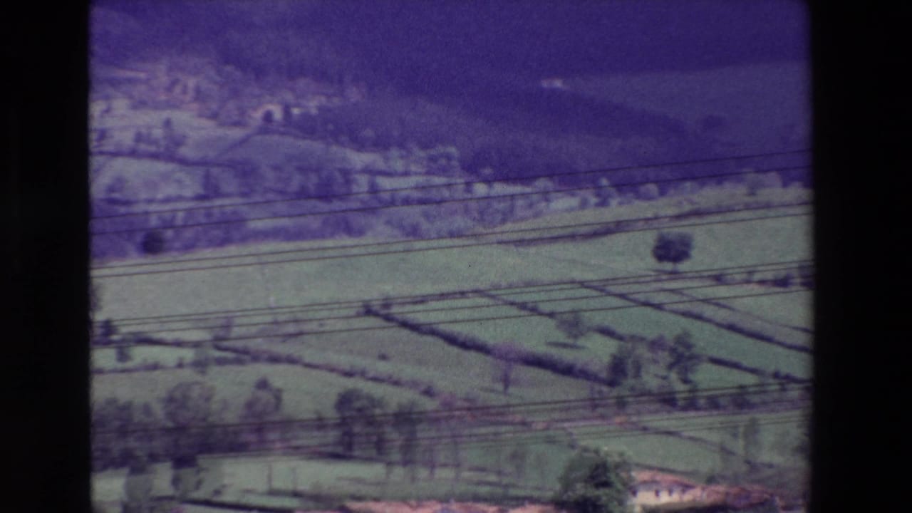 Soft 8mm frame showing green agricultural fields, hedgerow-like divisions, wooded slopes, a few small structures, and dark overhead lines across the view.