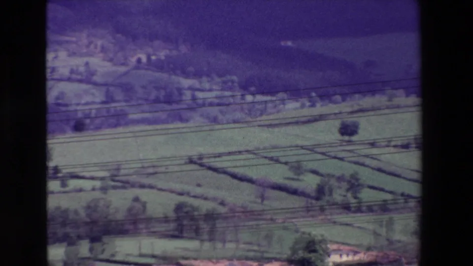 Soft 8mm frame showing green agricultural fields, hedgerow-like divisions, wooded slopes, a few small structures, and dark overhead lines across the view.