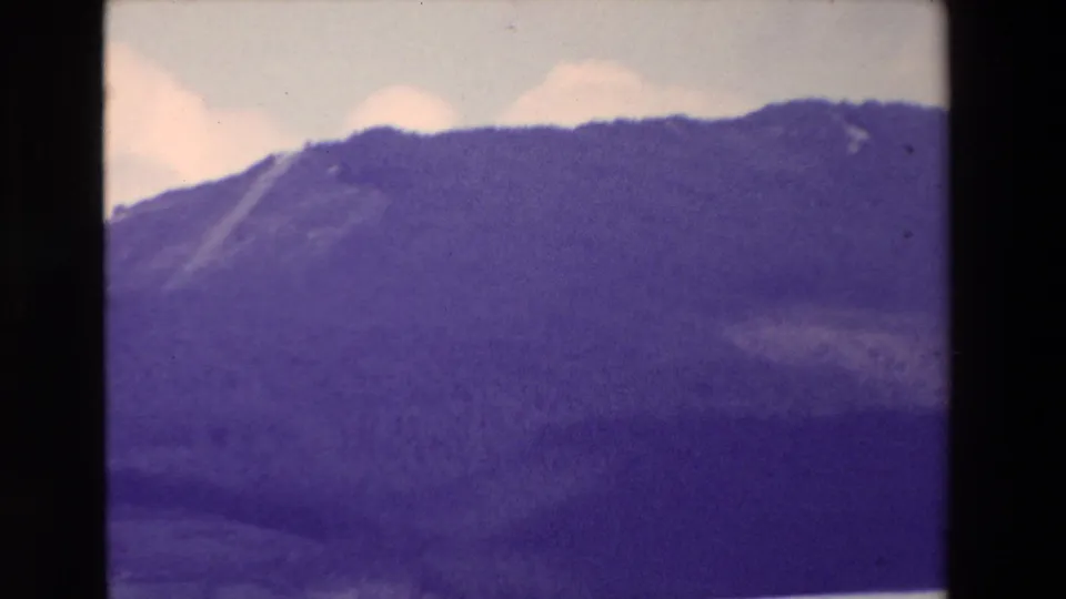 Home-movie frame with a dark mountain slope and pale clouds, the lower landscape softened by motion blur and purple color shift from the 8mm film scan.