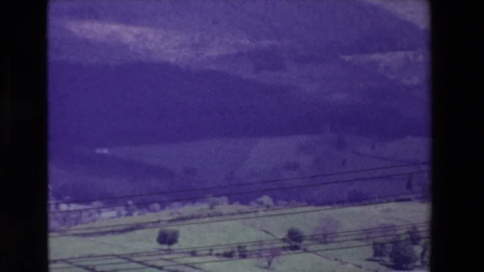 Late-1960s 8mm rural landscape frame with green fields, scattered trees, dark wooded hills, foreground utility lines, and pronounced purple-blue film color shift.