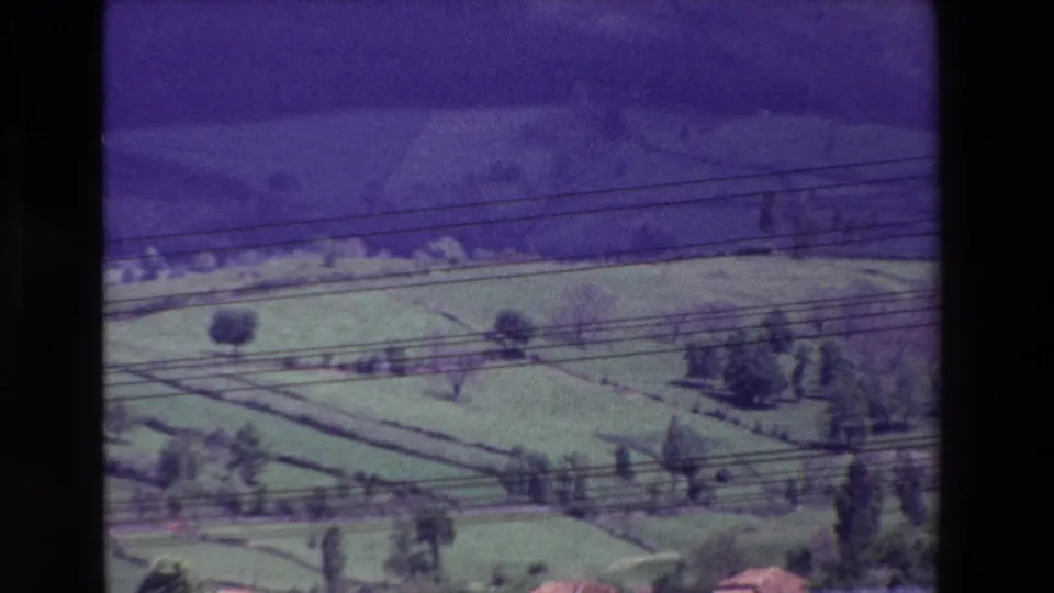 Soft 8mm frame of a rural hillside with terraced green fields, trees, and overhead power lines, with no people visible.