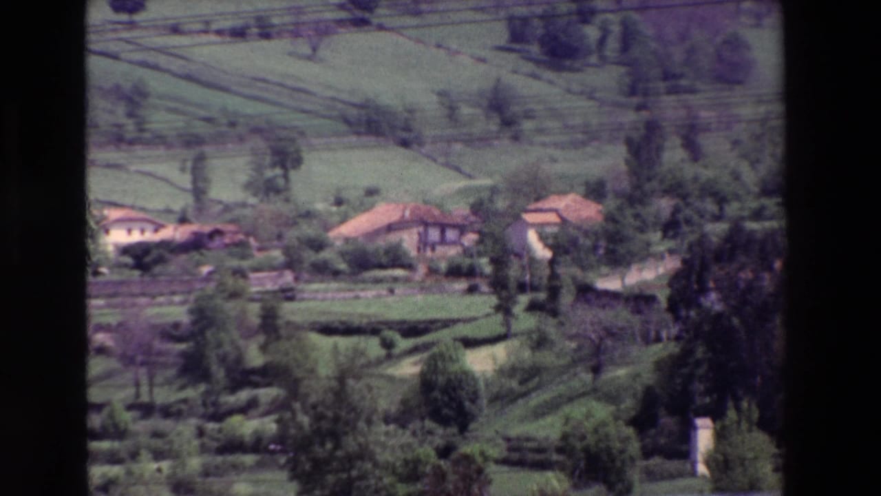 Vintage 8mm view of scattered rural houses with terracotta-colored roofs among green terraced fields and trees, with power lines visible across the upper frame.