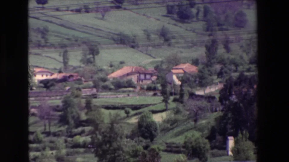Vintage 8mm view of scattered rural houses with terracotta-colored roofs among green terraced fields and trees, with power lines visible across the upper frame.