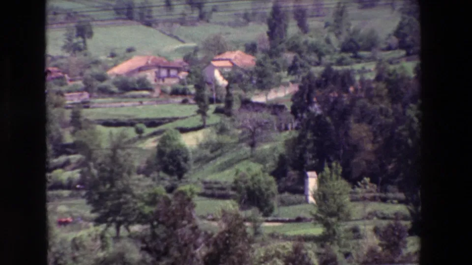 Home-movie frame showing rural houses, trees, and terraced farmland on a green hillside, with soft focus and visible film grain.