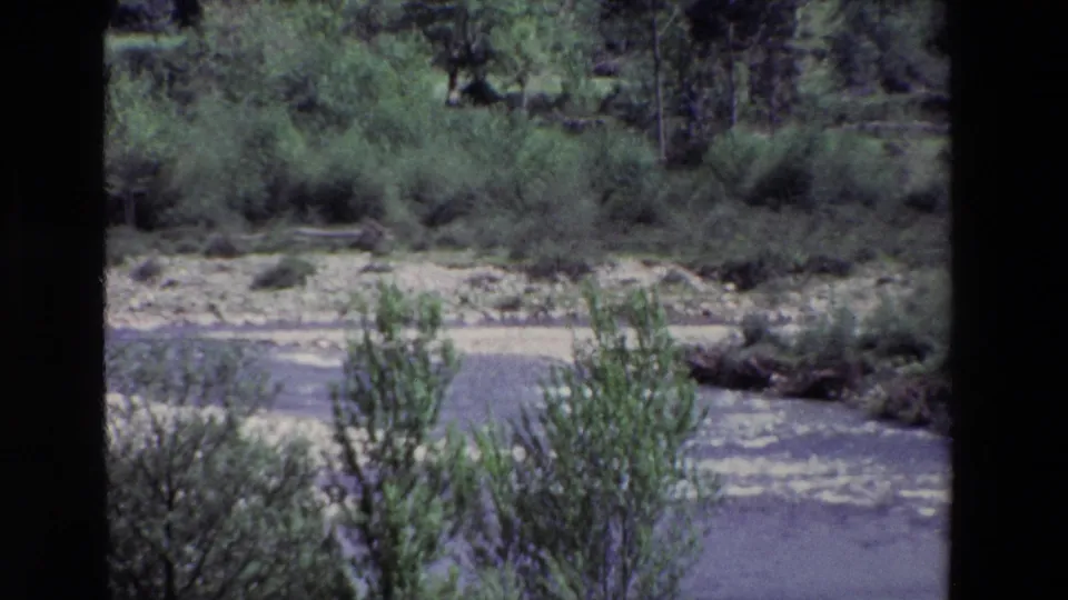 1969 archival 8mm footage from Spain showing a river bend with pale surface texture, low shrubs, trees, and exposed banks in a rural landscape.