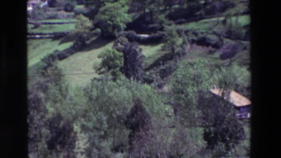 Archival 8mm frame of a green rural hillside, with trees and field boundaries surrounding a small farm structure in a Spanish countryside scene identified by the clip metadata.