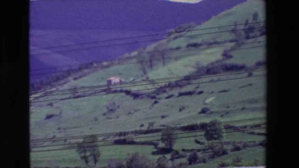 Archival 8mm wide view of terraced green hills and a small distant structure, with utility lines cutting across the rural landscape.