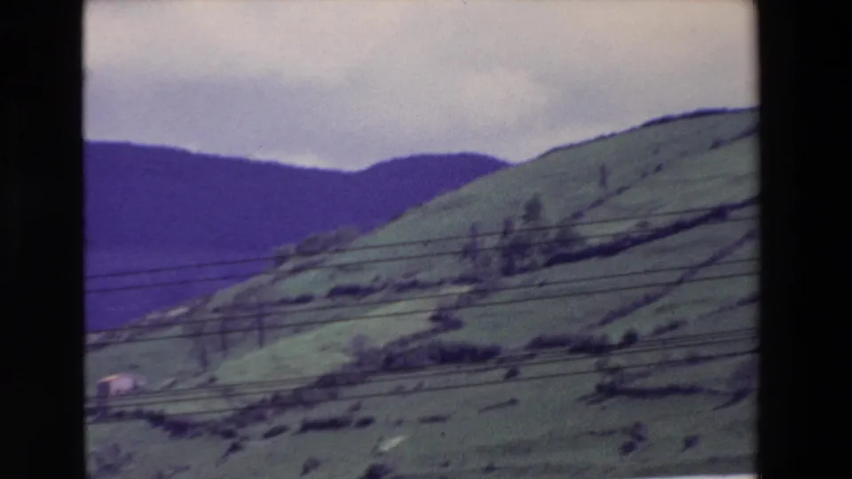 Archival 8mm frame of rolling green countryside with distant hills, overhead power lines, scattered trees, and a small building near the lower edge of the slope.